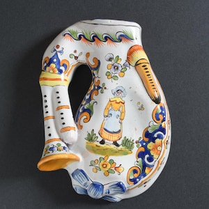 May include: A white ceramic wall pocket shaped like a musical instrument with a blue and yellow floral design. A small figure of a person in a blue and yellow dress is painted on the pocket.