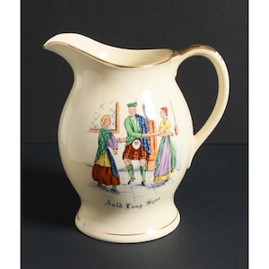 May include: Cream-colored ceramic pitcher with a gold rim and handle. The pitcher features a painted scene of three people in traditional Scottish attire, with the words "Auld Lang Syne" at the base.