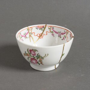 Könnte beinhalten: Weiße Porzellanschale mit floralen und Rankenmustern in Rosa, Grün und Gold. Die Schale wurde mit goldfarbenem Kintsugi, einer japanischen Kunstform, repariert. Die Schale hat einen kleinen Fuß.