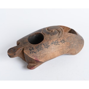 May include: A carved wooden turtle sculpture with a weathered brown finish. The shell features a circular swirl carving, a deep circular indentation, and black ink calligraphy characters. The turtle has a small head with visible eyes and mouth.