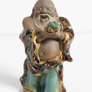 May include: A ceramic figurine of a smiling, bearded man holding a green and brown orb. The figurine is painted in shades of brown, green, and gold.