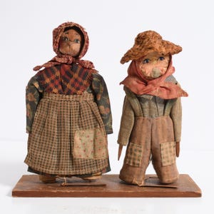 Può includere: Due bambole vintage di arte popolare, una donna e un uomo, in piedi su una base di legno. La bambola della donna indossa un vestito fantasia e un copricapo. La bambola dell'uomo indossa una tuta e un cappello. Entrambe le bambole hanno volti dipinti e sono fatte di tessuto.
