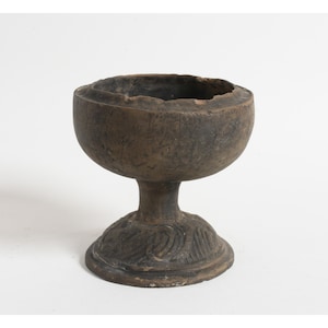 May include: A weathered, brown, chalice-shaped vessel with a wide bowl and a pedestal base. The base features a carved, swirling design. The vessel appears to be made of a dark, earthy material, possibly clay or stone.
