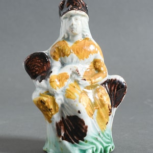 May include: A ceramic figurine of a seated woman with a hat. The figure is painted with white, yellow, brown, and green colors. The woman's hat is brown, and her dress has yellow and brown accents. The base is green.