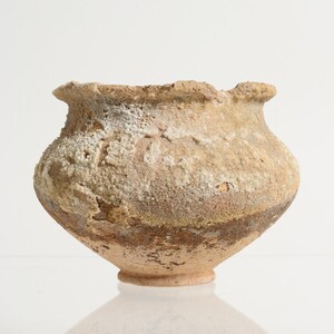 May include: A small, round ceramic pot with a wide, flared rim and a narrow base. The exterior surface features a rough, textured finish with mottled shades of beige, tan, and brown, accented by patches of white, crystalline mineral deposits.