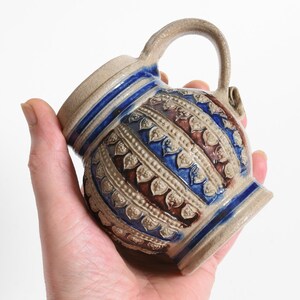May include: A small ceramic mug with a textured surface featuring alternating bands of cobalt blue and earthy brown glaze. The body is decorated with embossed leaf-like patterns and raised beaded lines, finished with a single curved handle.