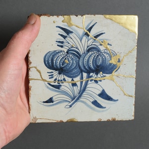 May include: A white ceramic tile with a blue and gold floral design. The tile is cracked and has a gold border.