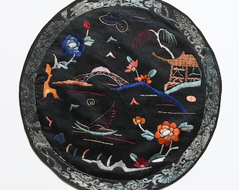 May include: Circular black fabric cushion with a silver border and colorful embroidered design of a landscape with a bridge, a pagoda, flowers, and a boat.