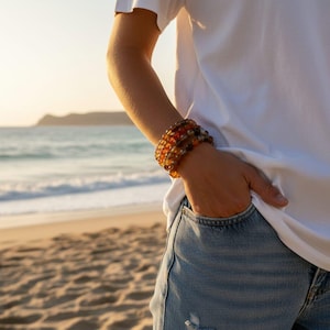 May include: A stack of amber and brown beaded bracelets. The bracelets are worn on a wrist, with a white shirt and blue jeans in the background. The beach and ocean are visible in the background.