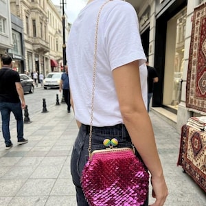 Puede incluir: Un bolso de mano fucsia con lentejuelas y una correa de cadena dorada. El bolso tiene un cierre de beso y está decorado con dos acentos esféricos coloridos. El bolso se lleva al hombro.
