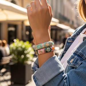 Puede incluir: Dos pulseras coloridas y ornamentadas están apiladas en una muñeca. La pulsera superior es turquesa con una piedra blanca, y la pulsera inferior es rosa y verde con elementos decorativos. La persona lleva una chaqueta vaquera.