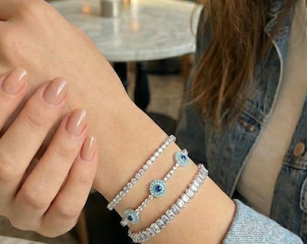 Puede incluir: Tres pulseras de plata adornan una muñeca. Una es una delicada hebra, otra presenta detalles azules en forma de ojo, y la tercera tiene piedras rectangulares. Las pulseras se llevan con una chaqueta vaquera.