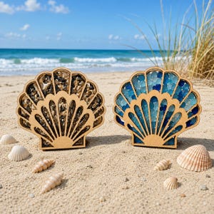 May include: Two shell-shaped wooden display frames with clear fronts. The left frame is filled with small, dark, triangular shark teeth. The right frame is filled with various shades of blue sea glass pieces. Both frames sit upright on the sand.