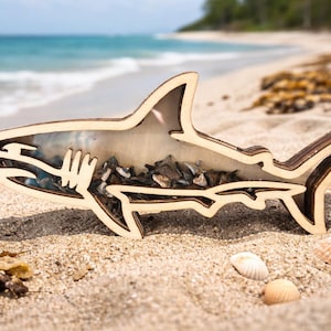 May include: A wooden shark ornament, light brown in colour, filled with small stones and shells. The shark is on a sandy beach with seashells. The background shows the sea and sky. The shark is approximately 30 cm long.