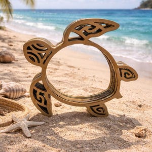 Peut inclure: Une sculpture de tortue en bois doré, composée de plusieurs couches avec des motifs découpés complexes. La tortue est posée sur une plage de sable avec une étoile de mer, des coquillages et l'océan en arrière-plan. La sculpture mesure environ 25 cm de long.