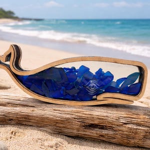 Könnte beinhalten: Ein dekoratives Wal-Objekt aus Holz, gefüllt mit blauem Meeresglas, auf Treibholz am Strand. Der Wal hat eine transparente Mitte mit Augendesign, das Meeresglas ist leuchtend blau.
