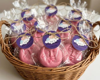 May include: A woven wicker basket filled with pink moon-shaped soaps individually wrapped in clear plastic bags. Each soap features a circular purple tag tied with twine, printed with a baby illustration and the text Eva's Baby Shower June 2024.