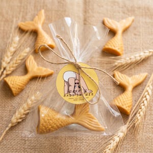 May include: A clear plastic bag containing a golden mermaid tail-shaped soap, tied with brown twine and a round yellow tag featuring a baby illustration and the text: SARAH'S BABY SHOWER THANK YOU FOR LOVING BABY SARAH JUNE 202X.