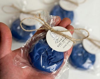 May include: A round, dark blue wax seal featuring a moon face design, individually wrapped in clear plastic with a tied jute twine bow and a circular white paper tag printed with a stork illustration and the text EVA'S BABY SHOWER JUNE 2026.