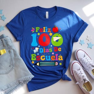 May include: A royal blue t-shirt with the words "Feliz 100 Dias De Escuela" in colourful letters and numbers. The design includes an apple, a paper aeroplane, a pencil, and stars. White trainers and denim shorts are also in the image.