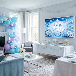 May include: A living room decorated for a birthday party with a blue and white theme. A large TV displays "Happy Birthday" in a wintery design. Balloons in shades of blue, silver, and purple adorn the room, along with a table set for a celebration.