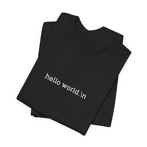 Op de afbeelding: Zwart gevouwen T-shirt met de witte tekst "hello world.
". De tekst is in een eenvoudige, schreefloze lettertype. De stof lijkt zacht.