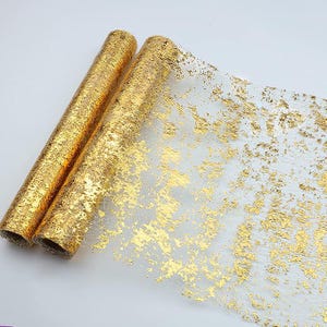 May include: A roll of sheer, translucent fabric featuring a metallic gold foil pattern. The material is tightly wound into a cylindrical shape on the left, while the unrolled portion on the right displays an irregular, textured gold leaf design.