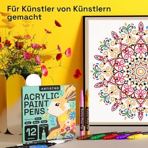 May include: A display of art supplies, including a box of ARTISTRO acrylic paint pens with a rabbit illustration, and a framed mandala artwork. The box indicates the pens are suitable for rocks, glass, wood, and paper. The image also shows colorful pens and a vase of flowers.