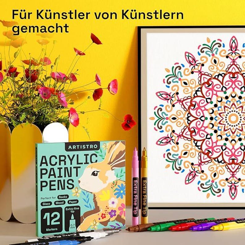 May include: A display of art supplies, including a box of ARTISTRO acrylic paint pens with a rabbit illustration, and a framed mandala artwork. The box indicates the pens are suitable for rocks, glass, wood, and paper. The image also shows colorful pens and a vase of flowers.
