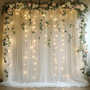 May include: A sheer white curtain backdrop adorned with string lights, faux flowers, and greenery. The floral arrangement includes white and pink roses. This decorative backdrop is suitable for weddings, parties, or photo shoots.