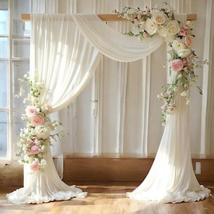 May include: A wooden wedding arch draped with flowing white fabric and adorned with floral arrangements of white and pink roses, peonies, and greenery. The arch is set against a backdrop of white curtains and a wooden floor, creating an elegant and romantic setting.