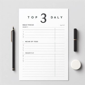 May include: A white daily planner sheet with the text "TOP 3 DALY" and lines for notes. A black pen with gold accents and a black pen are on the left and right. A white circular object is on the right.