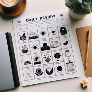 May include: A white "Daily Review" planner with black text and illustrations, including a sun, clouds, and flowers. The planner is on a white surface, next to a notebook, a pen, and a cup of coffee.