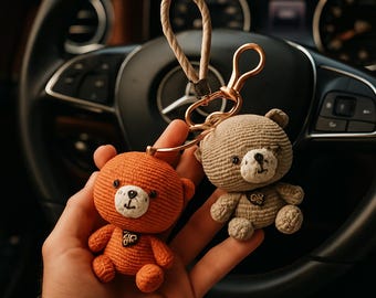 May include: Two crochet teddy bear keyrings. One is orange, the other is tan. Both have black eyes and a white muzzle. They are attached to a gold-coloured clasp and a beige rope loop. The background is a car's steering wheel.