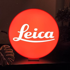 May include: A circular, illuminated sign featuring the white Leica logo centered on a vibrant red background. The sign emits a soft, warm glow, highlighting the brand name in its signature script font against the solid, circular red surface.