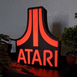 Puede incluir: Un letrero negro, texturizado y vertical que presenta el icónico logotipo de Atari y la palabra ATARI en letras mayúsculas negritas. El logotipo y el texto están iluminados con una luz roja brillante y vibrante, contrastando con la superficie mate.