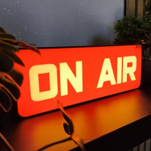 Può includere: Un'insegna rettangolare illuminata con uno sfondo rosso brillante e le parole ON AIR visualizzate in grassetto, in lettere maiuscole bianche. L'insegna emette una luce calda, evidenziando la tipografia chiara e senza grazie sulla superficie rossa.