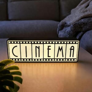 Può includere: Una scatola luminosa cinematografica rettangolare in bianco e nero con la parola "CINEMA" in maiuscolo. La scatola luminosa è su una superficie marrone chiaro, con un divano grigio sullo sfondo.