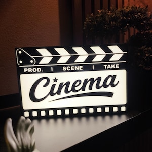 Peut inclure: Un panneau lumineux en forme de clap de cinéma, avec le mot "Cinema" écrit en gras. Le haut du clap est orné de rayures noires et blanches. Le panneau est posé sur une surface sombre.