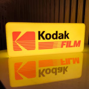 Può includere: Un cartello rettangolare giallo con il logo Kodak e la scritta «FILM» in arancione e nero. Il cartello è illuminato e si riflette su una superficie lucida.