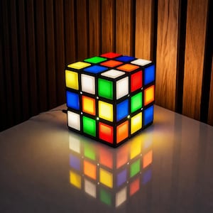 Könnte beinhalten: Eine würfelförmige Lampe, die an einen Rubik's Cube erinnert, mit einem schwarzen Rahmen und bunten Quadraten. Die Lampe leuchtet und zeigt Rot, Blau, Grün, Gelb, Orange und Weiß. Sie steht auf einer weißen Oberfläche und reflektiert die leuchtenden Farben.