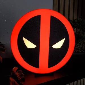 Könnte beinhalten: Eine kreisförmige Wandleuchte mit dem Deadpool-Logo, einem leuchtend roten Rand und einer schwarzen, strukturierten Mitte. Zwei leuchtend weiße, mandelförmige Augen befinden sich in der Mitte und bilden einen scharfen Kontrast zum dunklen Hintergrund.