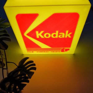 May include: A vintage Kodak sign with a yellow frame and a red and yellow logo. The sign reads "Kodak" and "FILM - CAMERAS - PRINTS". The sign is illuminated, casting a warm glow.
