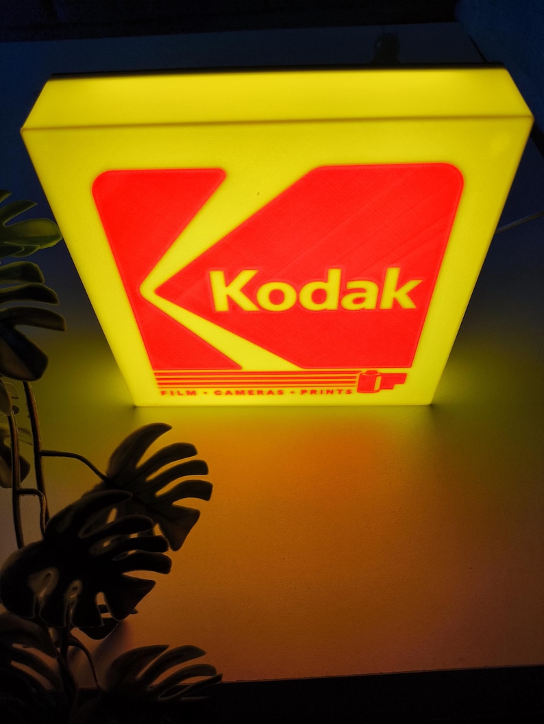 May include: A vintage Kodak sign with a yellow frame and a red and yellow logo. The sign reads "Kodak" and "FILM - CAMERAS - PRINTS". The sign is illuminated, casting a warm glow.
