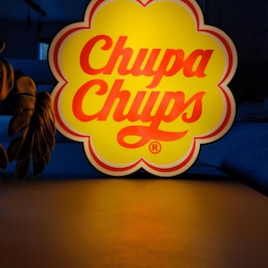 Könnte beinhalten: Ein Chupa Chups-Schild mit gelbem Hintergrund und roter Schrift. Das Schild hat die Form einer Blume und einen roten Rand. Das Chupa Chups-Logo ist deutlich zu sehen.