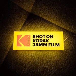 Może przedstawiać: Prostokątny żółty znak z logo Kodaka i czarnym napisem "SHOT ON KODAK 35MM FILM". Znak jest podświetlony, z czerwonym i pomarańczowym logo Kodaka.