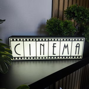 Könnte beinhalten: Ein rechteckiger Leuchtkasten mit einem schwarz-weißen Filmstreifen-Design und dem Wort CINEMA in fetten, schwarzen, serifenlosen Großbuchstaben. Der Leuchtkasten strahlt ein sanftes Licht aus, das das Muster des Filmstreifenrandes betont.