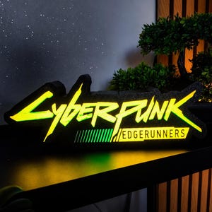 Könnte beinhalten: Ein schwarzes, beleuchtetes Schild mit dem gelb-grünen Schriftzug Cyberpunk Edgerunners. Das Schild hat einen strukturierten schwarzen Rand und strahlt ein helles Licht aus den Buchstaben aus, mit einer Reihe vertikaler grüner Linien unter dem Wort.