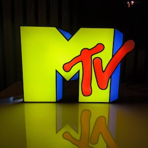 May include: Illuminated MTV logo in a studio setting. The large, three-dimensional sign features a bright yellow "M" with blue trim, and a red "TV" logo. The logo is reflected on the glossy surface below, creating a symmetrical image.