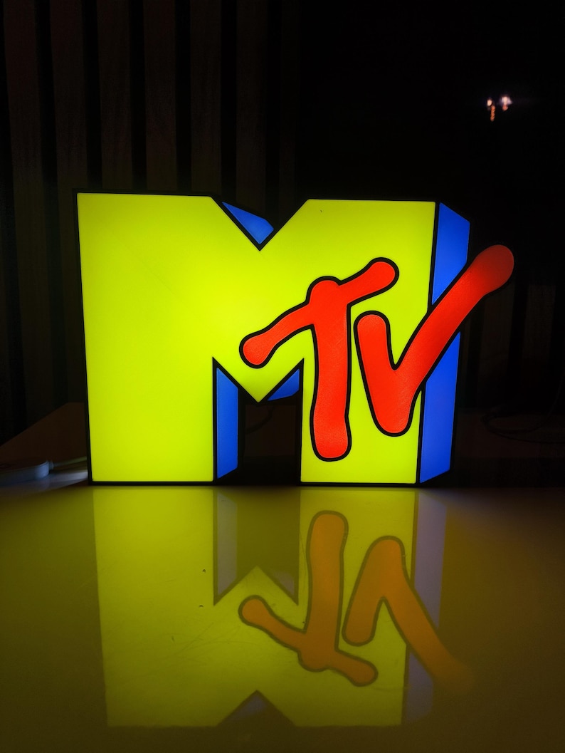May include: Illuminated MTV logo in a studio setting. The large, three-dimensional sign features a bright yellow "M" with blue trim, and a red "TV" logo. The logo is reflected on the glossy surface below, creating a symmetrical image.